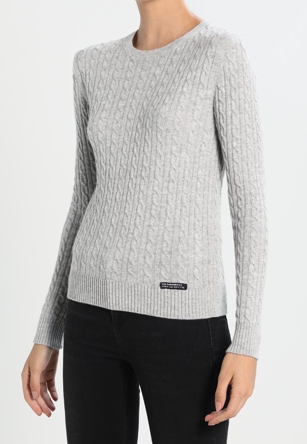 Femme portant un pull à torsades gris clair à manches longues et un pantalon noir, debout devant un fond blanc uni.