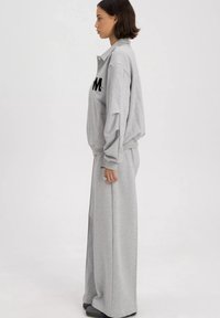 Grijze sweatshirt en brede broekset gemaakt van zachte stof. De sweatshirt heeft een zwart logo op de voorkant en heeft een kraag.