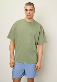 Olīvu zaļš pārāk liels T-krekls no mīksta kokvilnas auduma ar īsām piedurknēm un apaļu izgriezumu, kombinēts ar zilām svītrām biksēm.