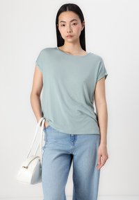 Lys grøn kortærmet top med løs pasform, parret med højdemmende lyseblå jeans og en hvid crossbody taske med guld lynlås detalje.