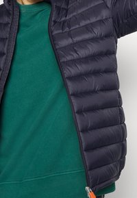 Granatowa kurtka puchowa z poziomymi przeszyciami i pełnym zamkiem błyskawicznym, nałożona na zielony sweter. Posiada kontrastującą pomarańczową zawieszkę do zamka.