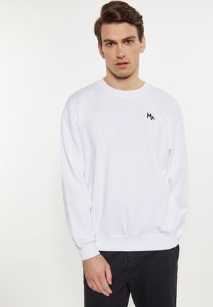 Ung mand iført en enkel hvid sweatshirt med teksten "Mr." på brystet og sorte bukser, stående mod en hvid baggrund.