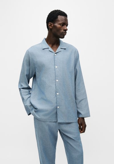 Homme portant une chemise bleu clair à manches longues boutonnée et un pantalon assorti, debout, une main dans la poche, regardant vers sa gauche.
