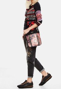 Woman wearing patterned sweater and black embroidered jeans, carrying a multicolored mandala-patterned crossbody bag, and black sneakers with brown laces.