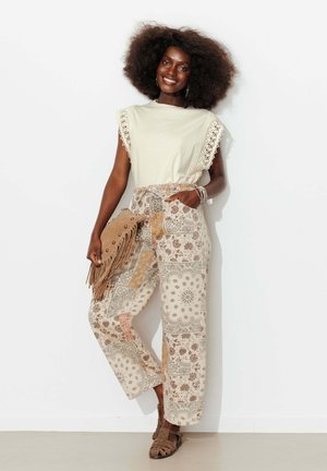 Femme aux grands cheveux bouclés portant un haut crème bordé de dentelle et un pantalon large à motifs, tenant une pochette marron à franges, souriante.