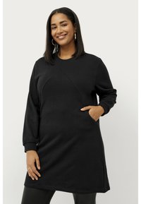Robe sweat noire avec des manches longues, un col rond, une poche latérale et un tissu texturé avec un détail de couture en diagonale.