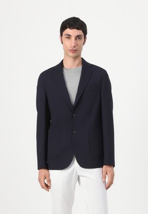 Homme portant un blazer bleu marine sur une chemise gris clair et un pantalon blanc, debout devant un fond clair uni.