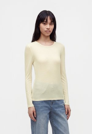 Junge Frau mit langen dunklen Haaren, die ein hellgelbes, langärmeliges, geripptes Top und blaue Jeans trägt, steht vor einem einfarbigen Hintergrund.