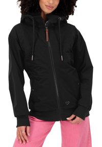 Donna che indossa una giacca nera con zip e cappuccio con lacci e pantaloni rosa, ritagliata dalle spalle in giù, in piedi contro uno sfondo bianco.