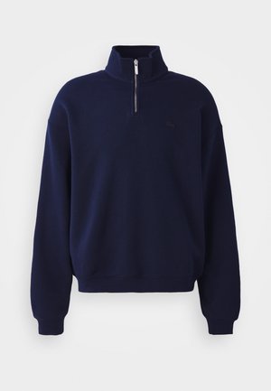 Donkerblauwe trui met lange mouwen, een halve ritskraag en geribde boorden, met een klein verhoogd geborduurd logo op de borst.