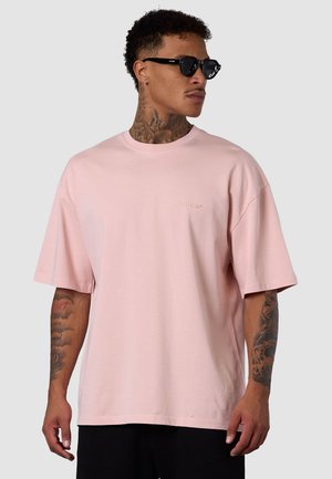 T-shirt surdimensionné rose clair en coton. Manches courtes, col ras du cou, et logo discret imprimé sur la poitrine. Texture lisse.