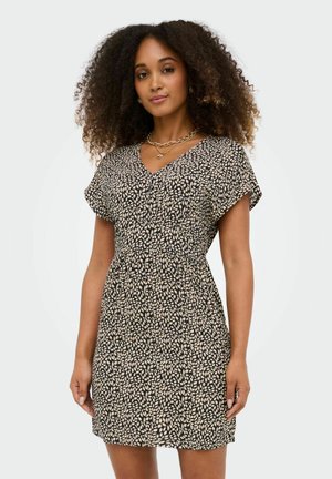 Göndör hajú nő fekete, rövid ujjú ruhában, krémszínű levélmintával, rétegezett arany nyakláncokkal kiegészítve.