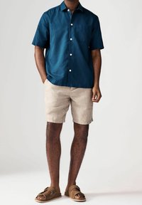 Camicia a maniche corte di colore verde scuro con bottoni, dotata di taschino, abbinata a pantaloni corti in lino beige chiaro e sandali scivolosi marroni.