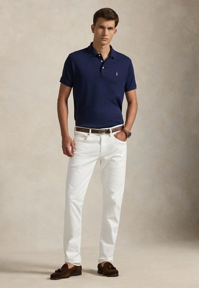 Junger Mann trägt ein marineblaues Poloshirt, weiße Hose, braunen Gürtel, braune Tassel-Loafer und eine Armbanduhr, steht mit einer Hand in der Tasche.