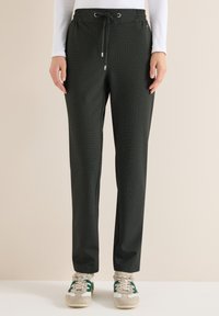 Person som bär mörka byxor med dragsko och vita sneakers med gröna ränder, stående mot en enkel ljus bakgrund.