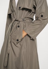 Trench coat khaki con vita cinturata, dettagli a bottoni e maniche lunghe. Tessuto liscio e design a strati, adatto per diverse occasioni.