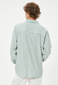 Camisa a rayas en azul claro y blanco, con cuello abotonado, mangas largas, dobladillo curvado, detalle de yugo en la parte posterior, hecha de tela suave.
