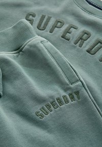 Bavlnená mikina a tepláky v vyblednutej zelenej farbe, s embosovaným logom "SUPERDRY", rebrovanými manžetami a mäkkou textúrou.