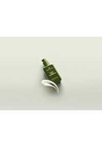 Groene, vierkante fles met een pompkop gelabeld "Origins," naast een gebogen witte applicator. Glad textuur, minimalistisch ontwerp, lichte achtergrond.