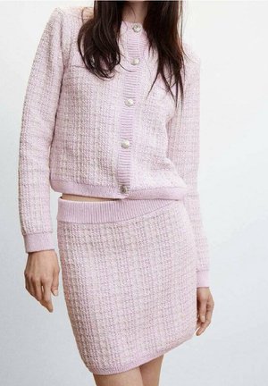 Femme portant un cardigan en maille texturée rose clair boutonné et une jupe taille haute assortie sur un fond blanc uni.