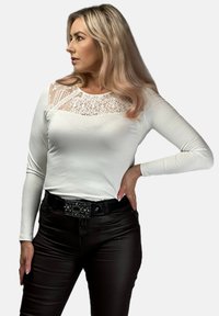 Biała bluzka z długim rękawem z koronkową wstawką na klatce piersiowej, zestawiona z ciemnobrązowymi dopasowanymi spodniami i ozdobnym czarnym paskiem.