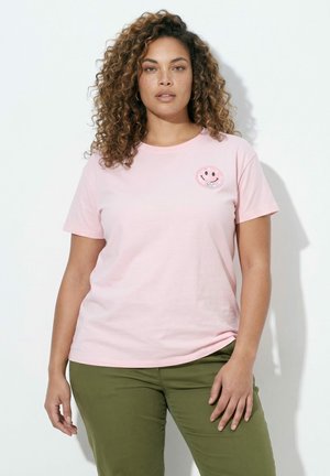 Woman with curly hair wearing a light pink T-shirt with a smiley face patch and olive green pants, standing against a white background.