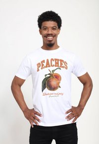 Vit bomullströja med ett tryck av persikor och texten "PEACHES" ovanför i orange och "Läckra och saftiga" nedanför i kursiv stil.