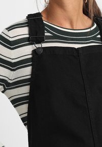Mustat farkkuhaalarit, joissa on säädettävät klipsihihnat, ovat päällä vihreä-kermanraidoitettu pitkähihaisessa paidassa. Tekstuuri näyttää tukevasti rakennetulta.