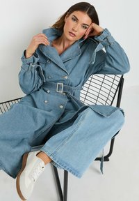 Jasnoniebieski denimowy płaszcz trench w podwójnym zapięciu, z dużym kołnierzem i paskiem w talii. Modelka ma na sobie białe sneakersy, siedząc na czarnej, drucianej krześle.