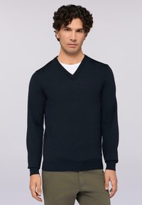 Navy-V-Ausschnitt-Pullover aus glatt gestricktem Material mit gerippten Bündchen und Saum. Getragen über einem weißen Hemd, kombiniert mit olivgrünen Hosen.