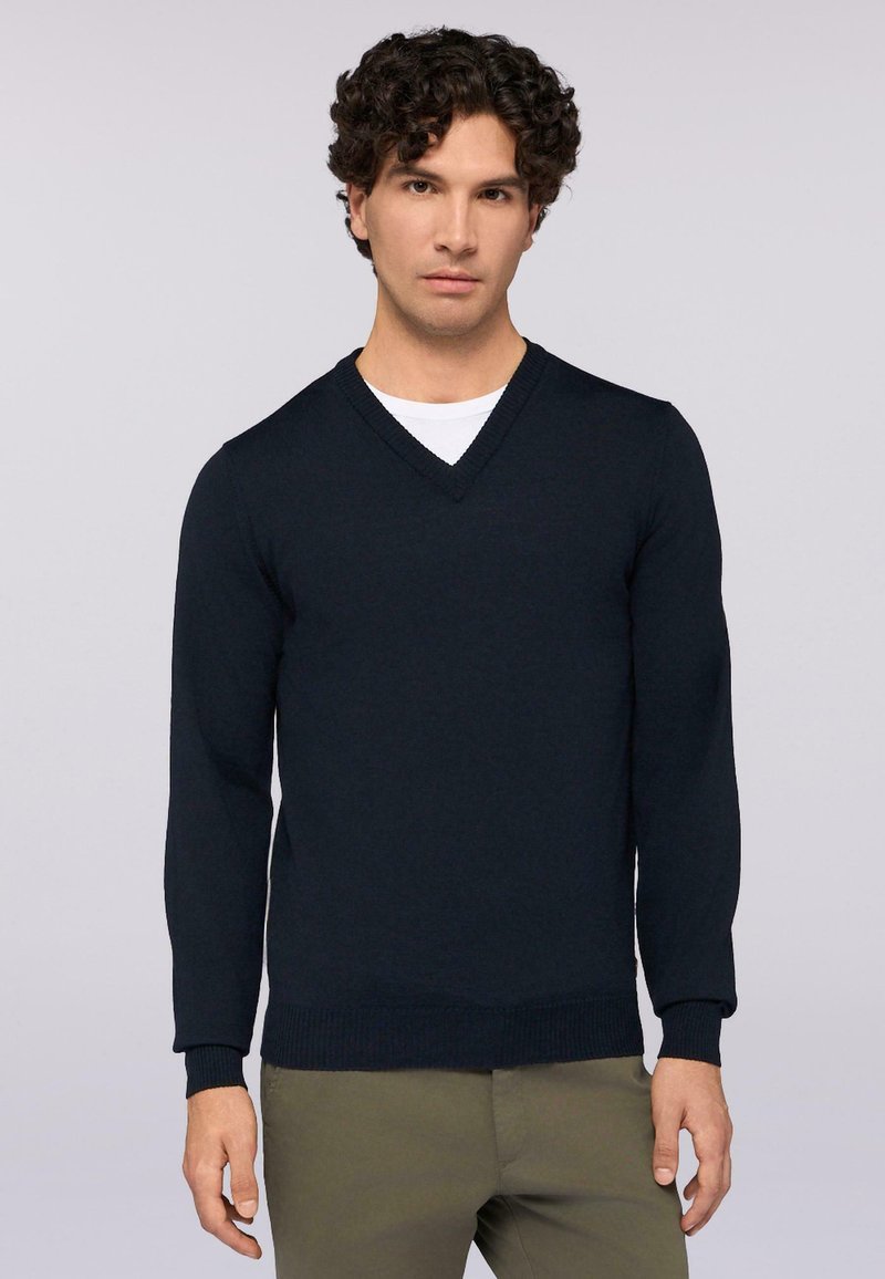 Navy-V-Ausschnitt-Pullover aus glatt gestricktem Material mit gerippten Bündchen und Saum. Getragen über einem weißen Hemd, kombiniert mit olivgrünen Hosen.