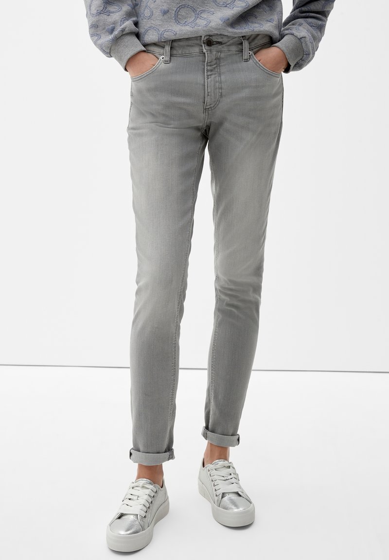 Personne portant un jean slim gris clair roulé aux chevilles et des baskets argentées et blanches, les mains dans les poches avant.