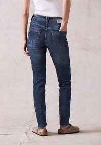 Jeans in denim blu scuro con vestibilità slim, vita media, due tasche posteriori e un leggero sbiadimento. Indossati con scarpe slip-on color beige.