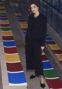 Svart skräddarsydd kavaj med fransdetaljer, matchande vida byxor och spetsiga skor, framför färgglada läktare.