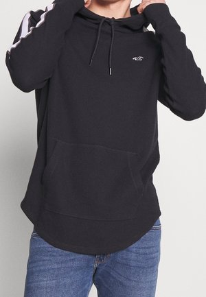 Osoba poprawiająca czarną bluzę z kapturem z białymi paskami na rękawach, kieszenią kangurka i małym białym logo na piersi, ubrana w niebieskie dżinsy.