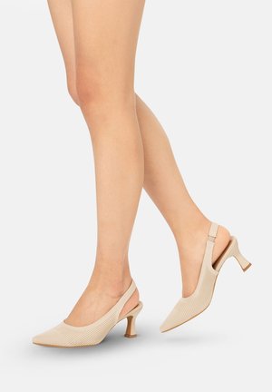 Benen met beige slingback kitten heels met een gestructureerd bovenwerk, getoond tegen een effen witte achtergrond.