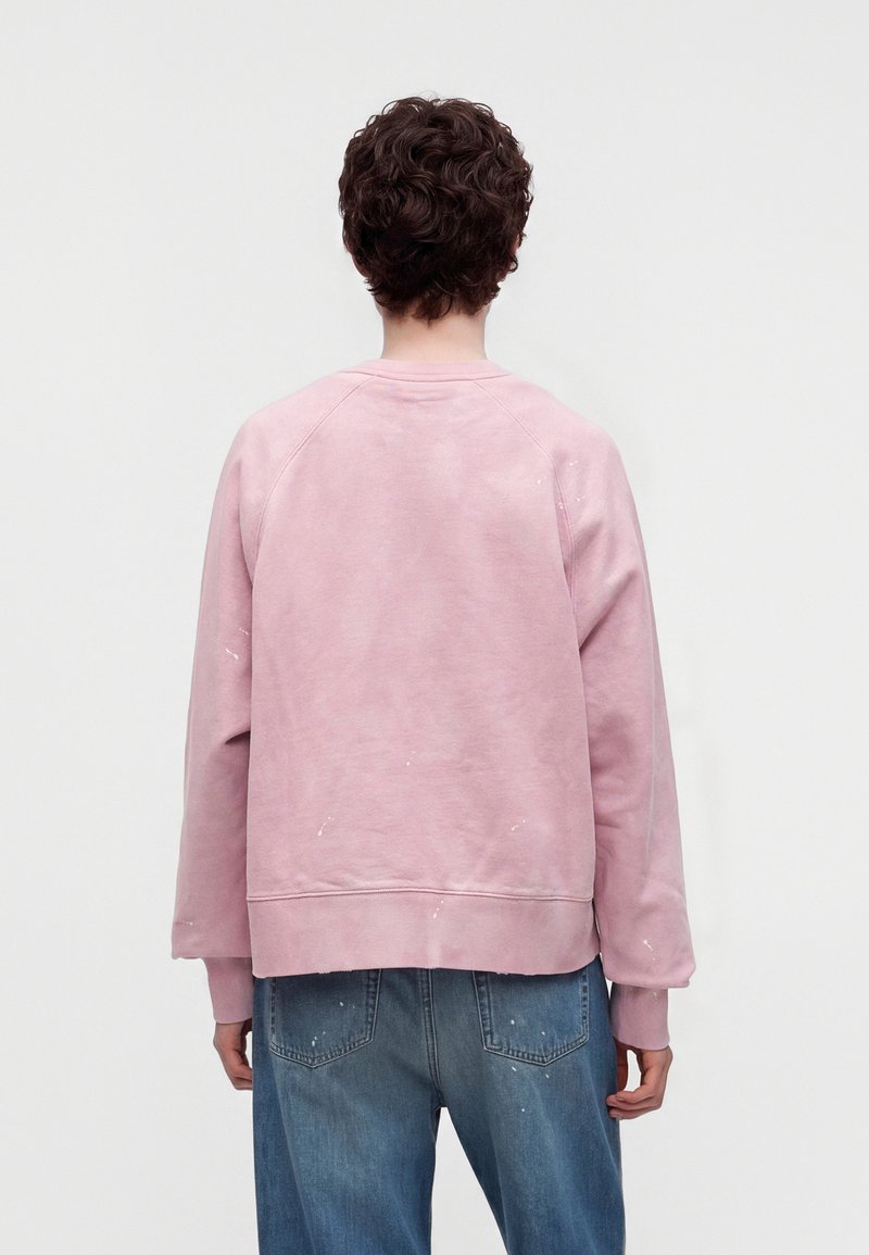 Person mit kurzen lockigen dunklen Haaren, die einen staubig rosa Sweatshirt und blaue Jeans mit Farbspritzern trägt, mit dem Rücken zu einem schlichten hellen Hintergrund.