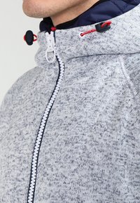 Närbild av en person som bär en gråspräcklig zip-up hoodie med en marinblå huva och röda dragsnören.