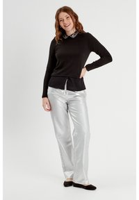 Black long-sleeve top with a jeweled collar, paired with shiny silver wide-leg pants. Model stands with one hand on hip, wearing black flats.