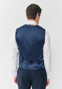 Marineblaues Satin-Weste mit floralem Muster am Kragen, hinten mit Verschluss, glatte Textur und verstellbarem Details des Gürtels, getragen über einem weißen Hemd.
