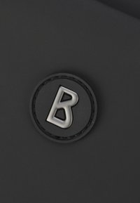 Accessorio nero con un'etichetta nera circolare in rilievo recante una "B" argentata al centro, su uno sfondo a texture liscia.
