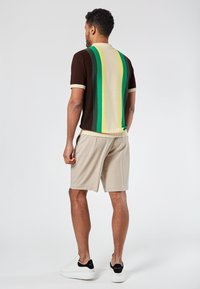 Camisa de polo de punto marrón con rayas verticales en amarillo, verde y crema, combinada con pantalones cortos beige y zapatillas blancas, vista desde la parte de atrás.