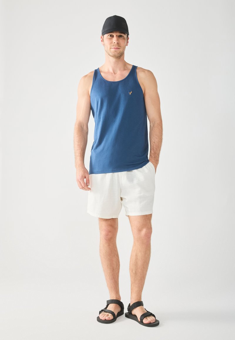 Canotta di cotone blu con un piccolo logo, pantaloni corti di lino bianchi e sandali neri. Il modello è in piedi contro uno sfondo neutro.