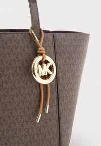 Torba tote iz rjave teksturirane tkanine z geometrijskim vzorcem, z zlatim kovinskim okrasjem in rjavim usnjenim dodatkom s krožnim logotipom.