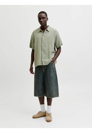 Joven con camisa verde claro de manga corta abotonada, pantalones cortos de mezclilla anchos y oscuros, calcetines blancos y zapatos beige, de pie sobre un fondo liso.