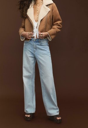 Kobieta w brązowej kurtce z owczej skóry noszona na biały top z wiązaniem z przodu, jasnoniebieskie dżinsy z szerokimi nogawkami, czarne sandały na platformie oraz warstwowa złota biżuteria.