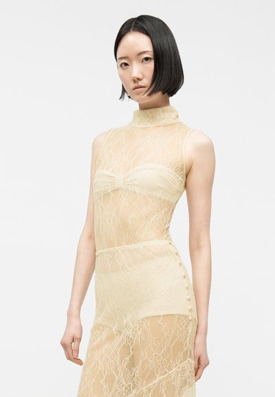 Νεαρή γυναίκα με ίσιο μαύρο καρέ χτένισμα φοράει αμάνικο μπεζ φόρεμα από δαντέλα και στέκεται μπροστά σε απλό ανοιχτόχρωμο φόντο.