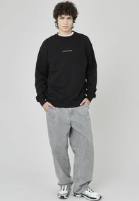 Sweatshirt noir avec un col rond et un texte blanc. Pantalons gris amples au fini texturé. Personne se tient avec les mains dans les poches.