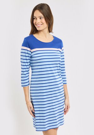 Femme souriante, portant une robe bleue rayée avec des manches trois-quarts, debout devant un fond uni gris clair.