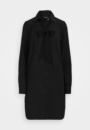 Robe-chemise noire à manches longues avec boutons sur le devant, plastron plissé sur la poitrine, col et grand nœud noué au niveau de l'encolure.
